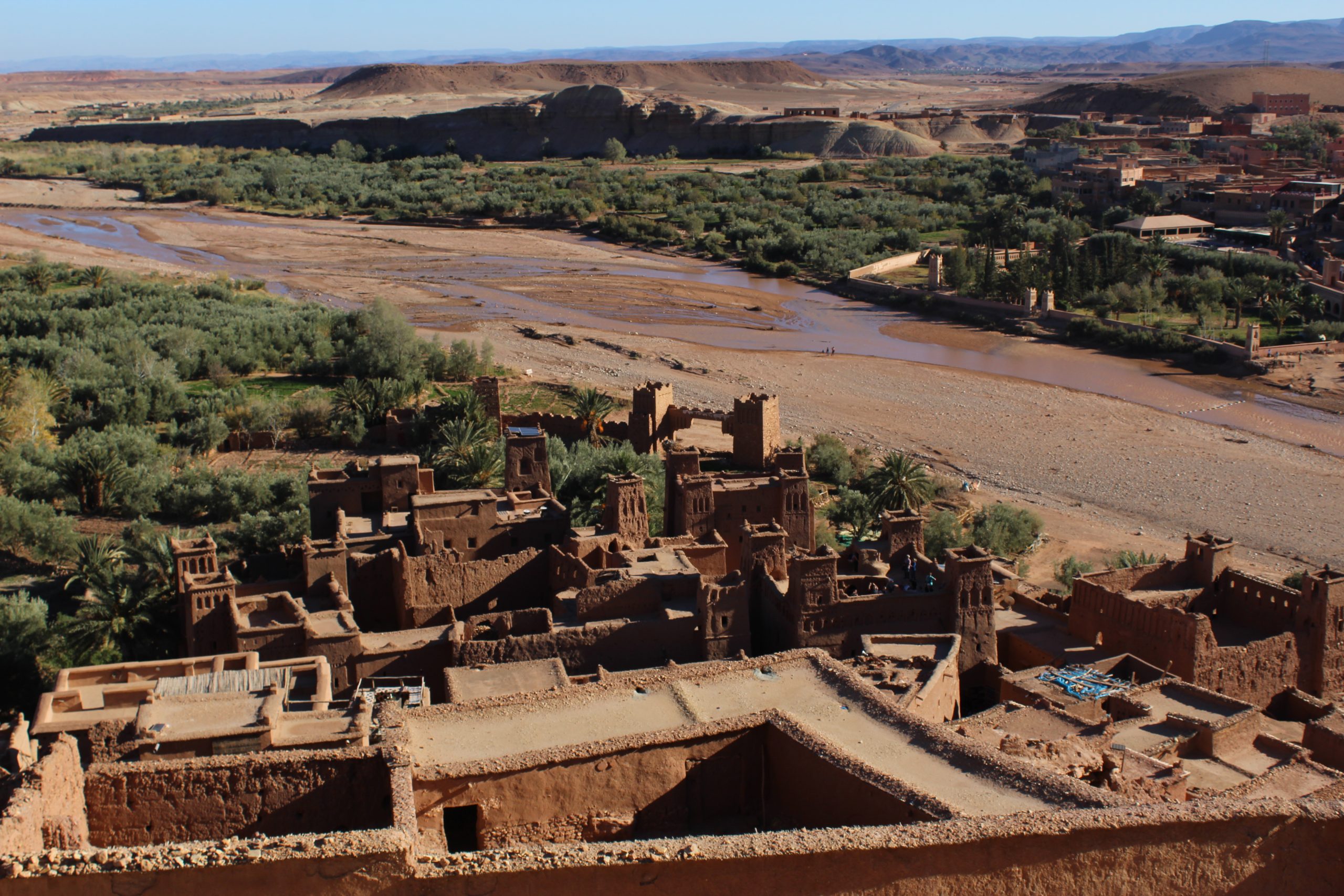 Aerial view of Aït Ben Haddou in Morocco by a dry riverbed, surrounded by green vegetation.